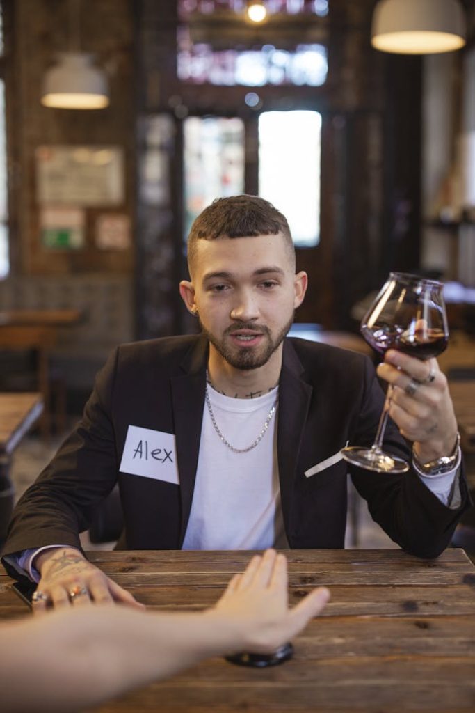 Man in black blazer at speed dating, holding wine glass, with name tag in café setting.